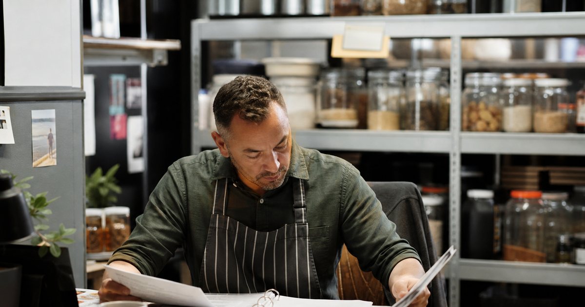 Restaurant owner at a small office desk in the back of house with a paper binder, printed recipe sheets, a calculator and a coffee mug, shelves with dry goods behind