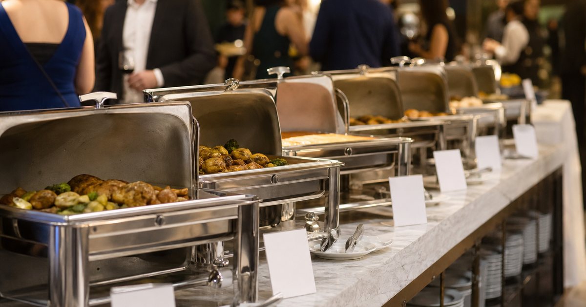 Elegant hotel buffet line with chafing dishes, small blank tent cards in front of each dish, tongs resting on saucers, guests in the background at an evening event