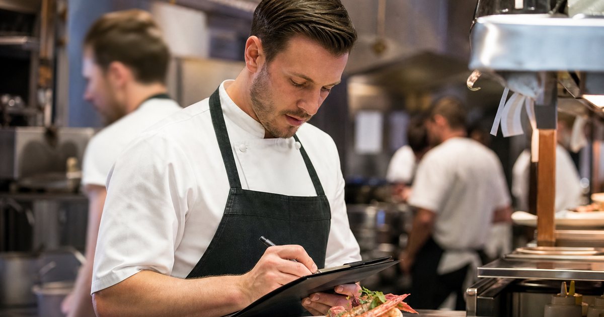 Chef writing on a clipboard next to a plated seafood special on a stainless pass in a busy restaurant kitchen, ticket rail, warm lighting, cooks in background