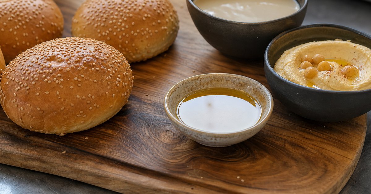 Close-up of a wooden board with sesame seed burger buns, a bowl of tahini, hummus and a small dish of sesame oil on a restaurant prep counter, soft daylight